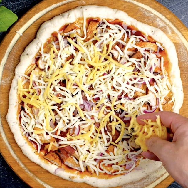 Cheese being sprinkled on BBQ chicken pizza before popping it into the oven.