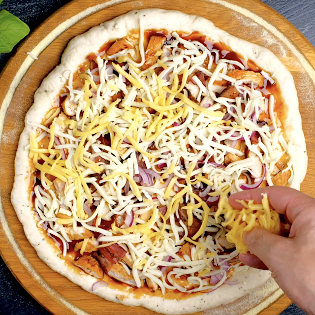 Cheese being sprinkled on BBQ chicken pizza before popping it into the oven.