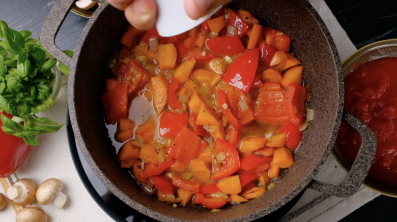 A pot with tomato, bell pepper, carrot mixture in liquid that's being seasoned with salt.