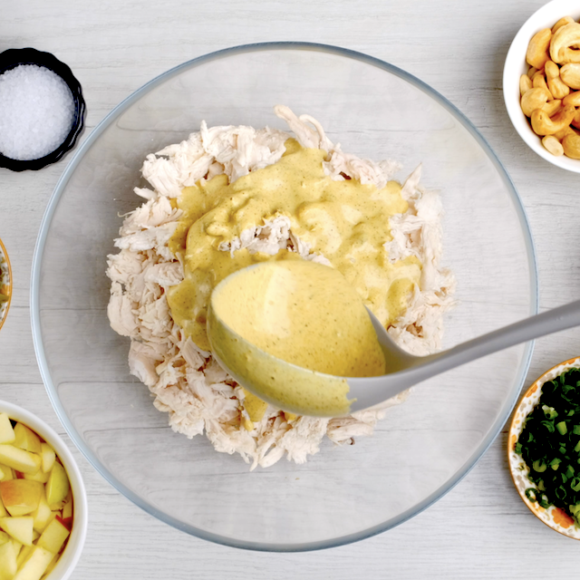 Curry dressing being ladled over shredded cooked chicken to make chicken curry salad at home.