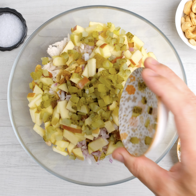 Chopped pickles, onions, apples, and raisins being added to bowl to make chicken curry salad.