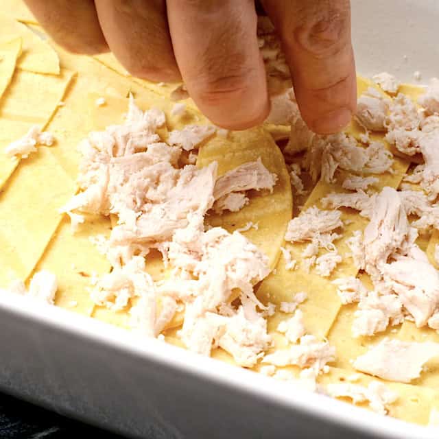 Shredded chicken being added on top of a layer of tortilla and soup mixture to make white chicken enchilada casserole.