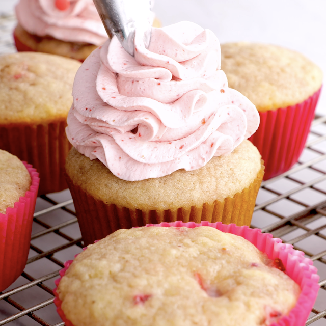 Strawberry cupcakes being piped with whipped cream frosting.