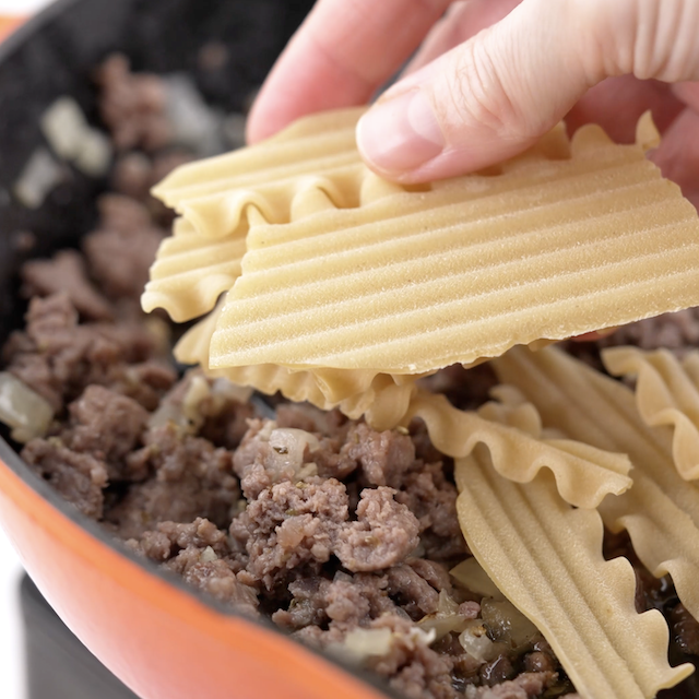 Broken lasagna noodles being added to a pan with cooked sausage meat.