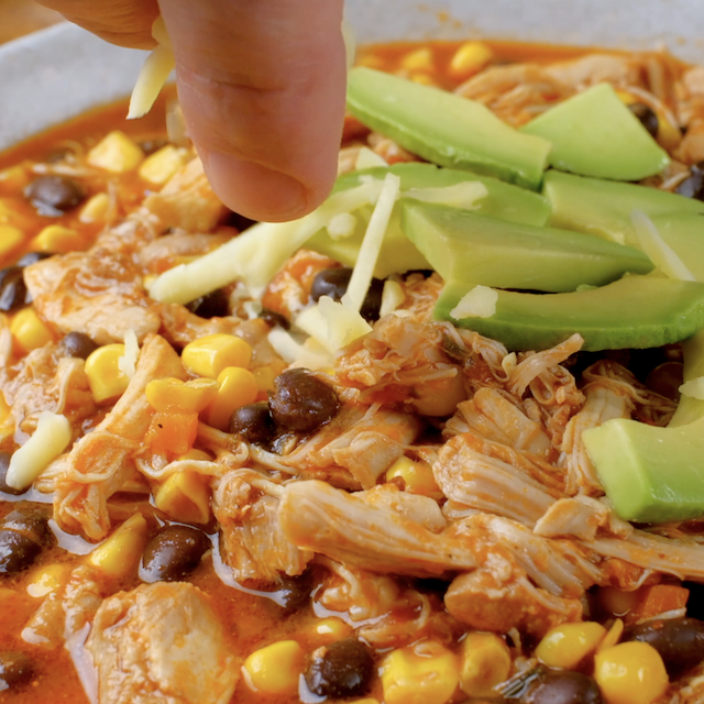 Freshly made crockpot salsa chicken being topped with sliced avocado and shredded cheese before serving.
