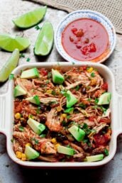 Crockpot chicken salsa in a casserole dish with avocado chunks on top with salsa on the side and lime wedges.