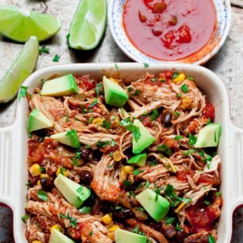 Crockpot chicken salsa in a casserole dish with avocado chunks on top with salsa on the side and lime wedges.