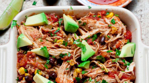Crockpot chicken salsa in a casserole dish with avocado chunks on top with salsa on the side and lime wedges.
