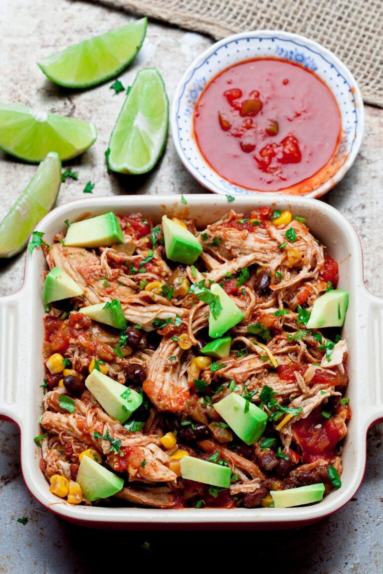 Crockpot chicken salsa in a casserole dish with avocado chunks on top with salsa on the side and lime wedges.