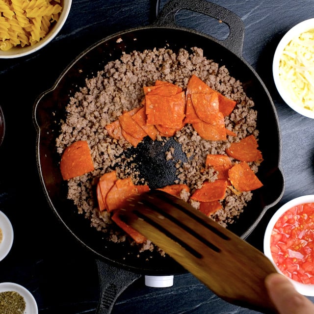 A skillet with browned ground sausage and pepperoni slices being stirred with a wooden spatula. 