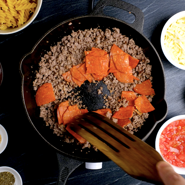 A skillet with browned ground sausage and pepperoni slices being stirred with a wooden spatula. 