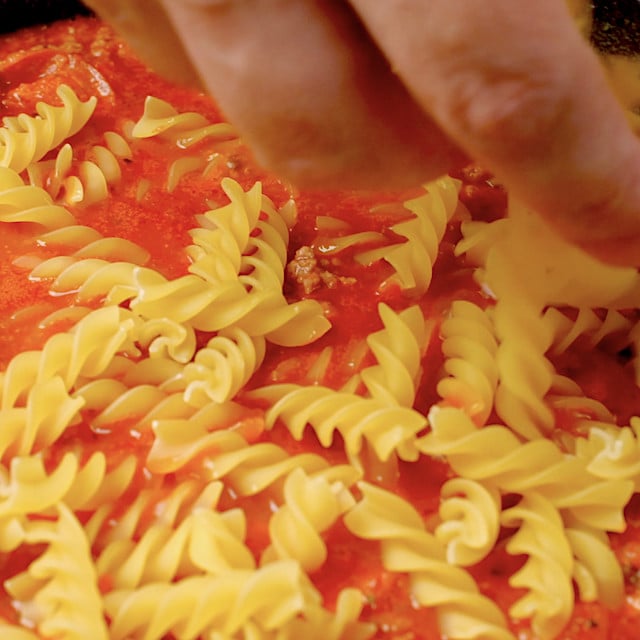 Uncooked pasta being added to pizza pasta sauce in a pot.
