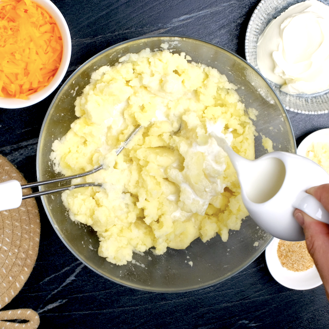 Milk being poured into mashed potatoes in a glass bowl.