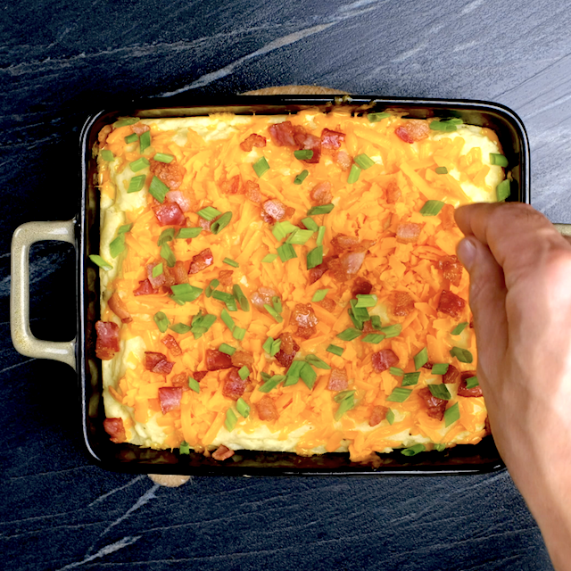 Creamy mashed potatoes being garnished with bacon, cheese, and green onions before baking.