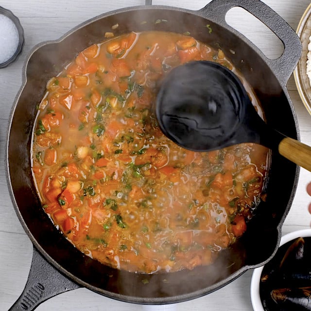 Hot broth being added to tomato and herb mixture in a pan to make easy seafood paella.