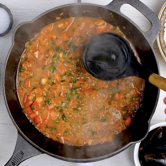 Hot broth being added to tomato and herb mixture in a pan to make easy seafood paella.