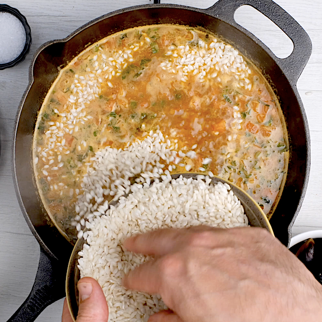 Rice being sprinkled over paella mixture in a hot pan.