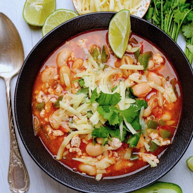 white bean turkey chili in a black bowl with sliced limes and cheese and cilantro. 
