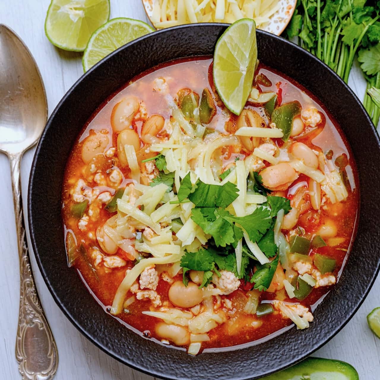 white bean turkey chili in a black bowl with sliced limes and cheese and cilantro.