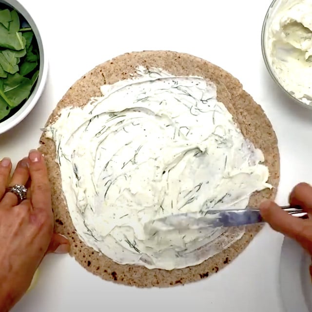 Creamy lemon dill spread being spread on a tortilla to make smoked salmon pinwheels.