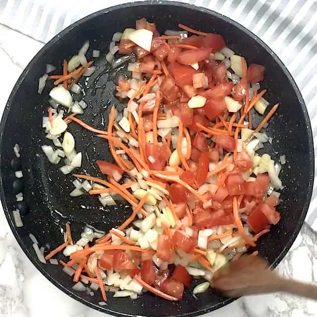 Chopped onions, carrots, tomatoes, and garlic are being sautéed in a black frying pan with a wooden spoon to make flavorful Thai fried rice.