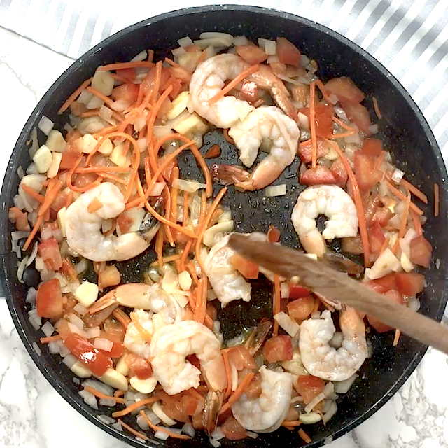 Shrimp, sliced carrots, tomatoes, and chopped garlic are being sautéed in a black frying pan with a wooden spoon, creating the flavorful base for Thai fried rice.