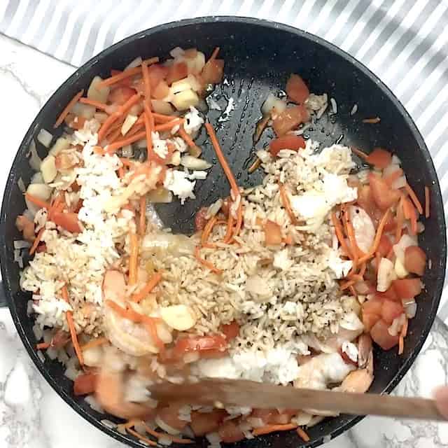 A pan with cooked rice, shrimp, chopped tomatoes, carrots, onions, and soy sauce being stirred with a wooden spoon for a delicious quick fried rice.
