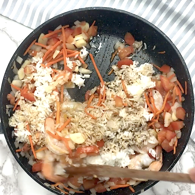 A pan with cooked rice, shrimp, chopped tomatoes, carrots, onions, and soy sauce being stirred with a wooden spoon for a delicious quick fried rice.