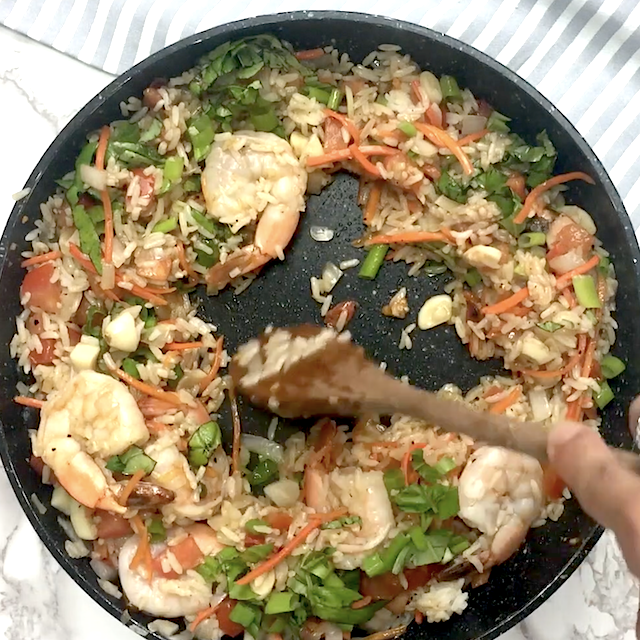 A person stirs Thai fried rice with shrimp, vegetables, and garlic in a black skillet using a wooden spoon.