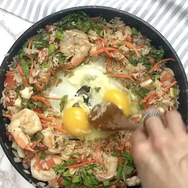 A hand stirs two raw eggs into a pan of shrimp, rice, carrots, green onions, and other vegetables on a stovetop, creating a delicious Thai fried rice.