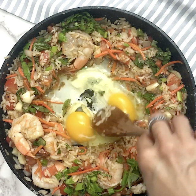 A hand stirs two raw eggs into a pan of shrimp, rice, carrots, green onions, and other vegetables on a stovetop, creating a delicious Thai fried rice.