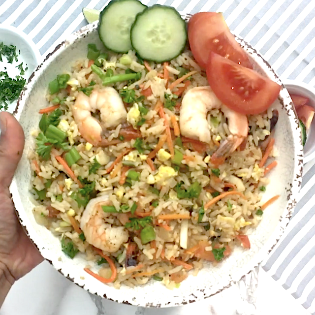 A plate of Thai fried rice with shrimp, garnished with chopped green onions, cucumber slices, and tomato wedges, held above a striped surface.