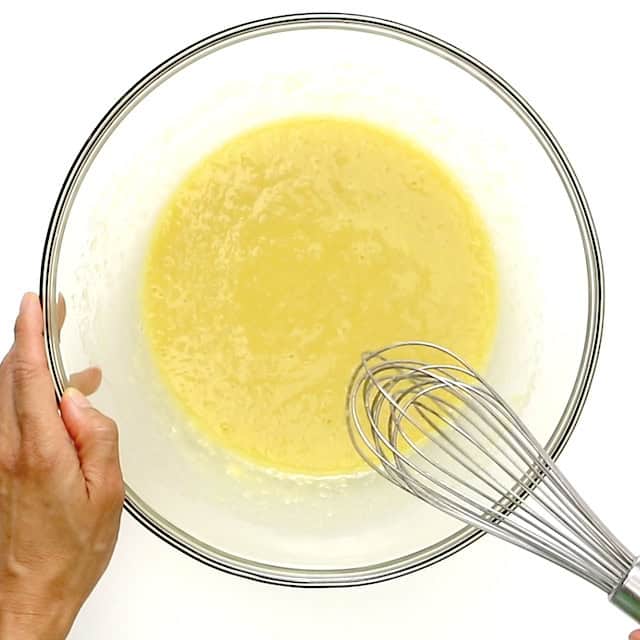 A hand holding a glass mixing bowl with yellow batter being whisked using a metal whisk.