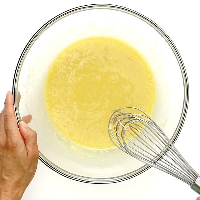 A hand holding a glass mixing bowl with yellow batter being whisked using a metal whisk.