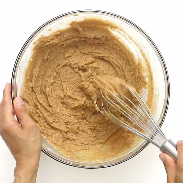 A person mixing a light brown spiced cake batter in a glass bowl with a metal whisk, viewed from above against a white background.