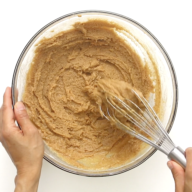 A person mixing a light brown spiced cake batter in a glass bowl with a metal whisk, viewed from above against a white background.