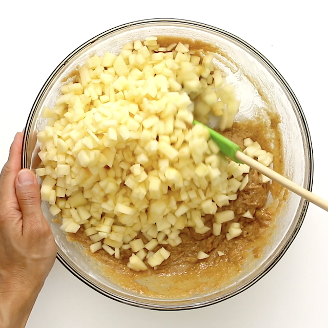 A hand mixes chopped apples into a bowl of batter using a green spatula, preparing an easy apple cake.