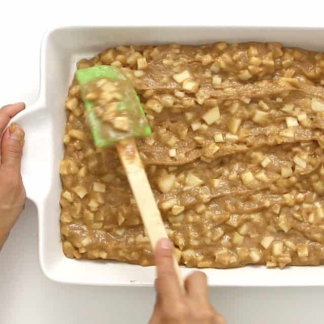 A person spreads easy apple cake batter with a green spatula in a white rectangular baking dish.