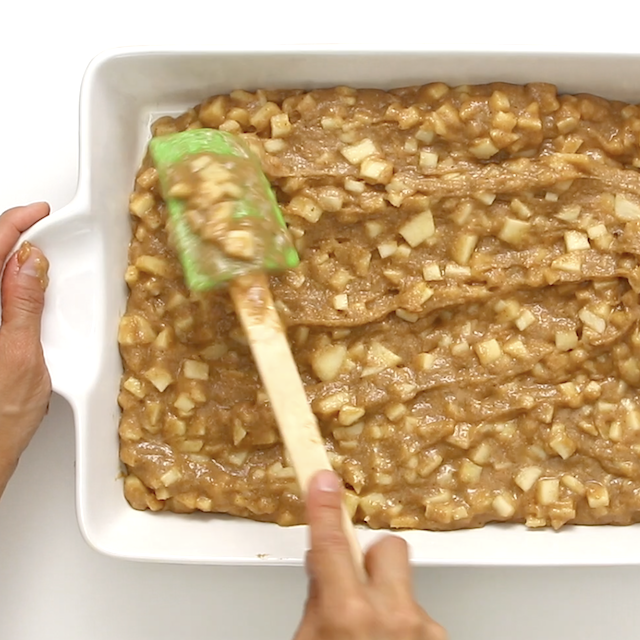 A person spreads easy apple cake batter with a green spatula in a white rectangular baking dish.