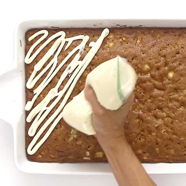 Cream cheese frosting being piped in diagonal lines onto a large rectangular apple cake.