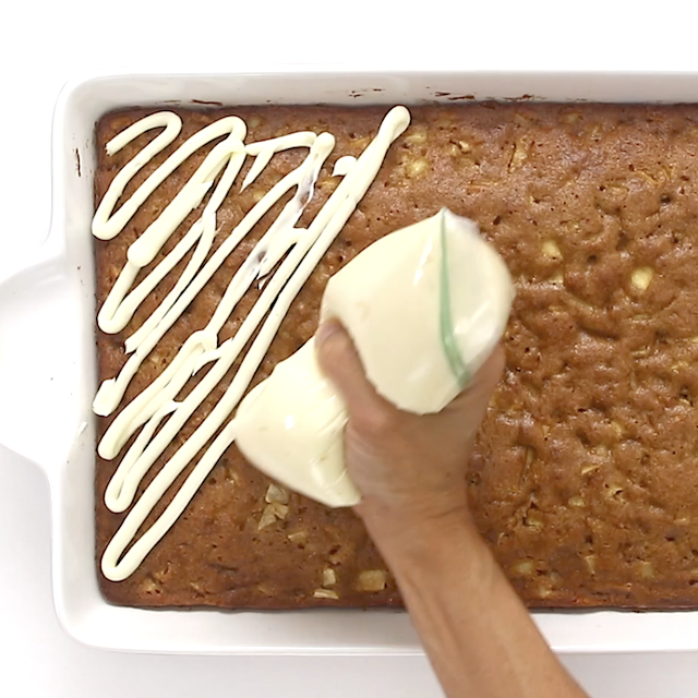Cream cheese frosting being piped in diagonal lines onto a large rectangular apple cake.