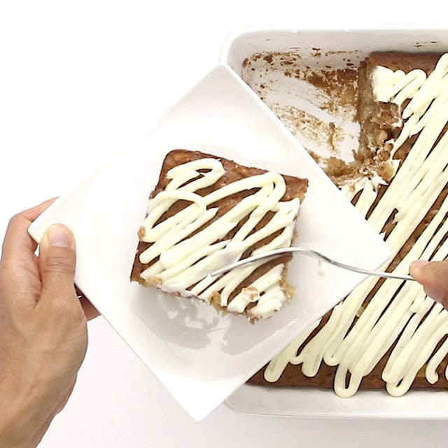 A person holds a square piece of easy apple cake on a plate with a fork, next to a baking dish containing the rest of the cake.