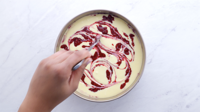A person swirling raspberry sauce into cheesecake mixture in a springform pan.