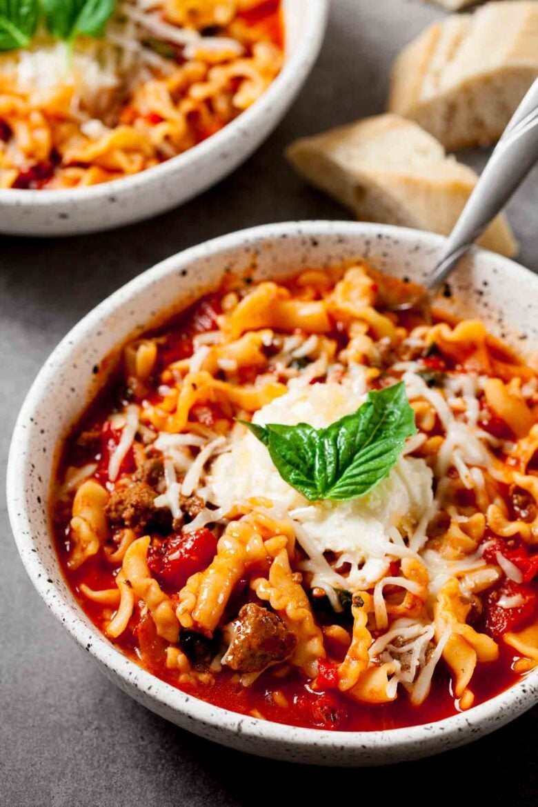 A bowl of Easy Lasagna Soup Recipe topped with shredded cheese and a fresh basil leaf, served with pieces of bread and another bowl of soup in the background.