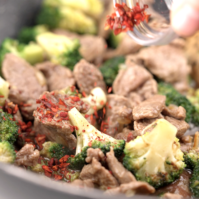Red crushed pepper being sprinkled over beef and broccoli stir fry before serving.