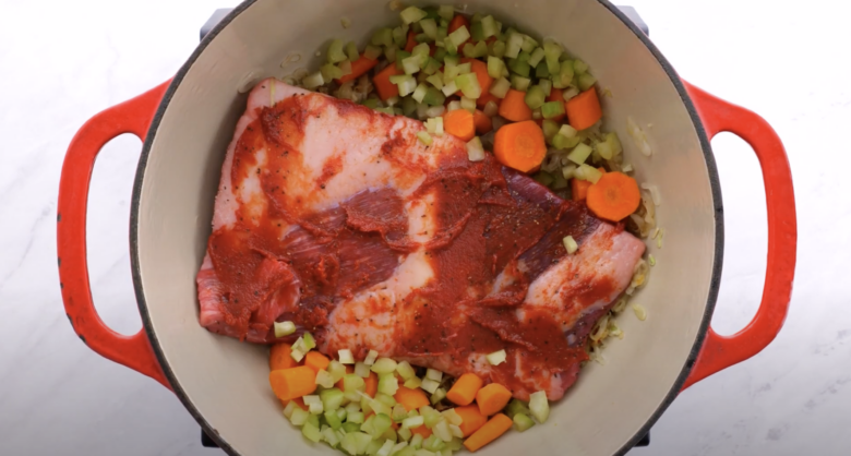 Seasoned brisket with tomato paste on top, surrounded by chopped vegetables in a Dutch oven.