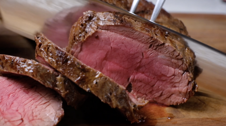 Cooked beef tenderloin being sliced on a cutting board.