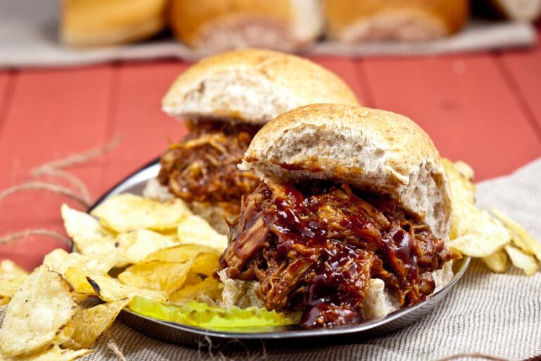 Pulled pork sandwiches with barbecue sauce on a plate, accompanied by potato chips and a pickle slice, placed on a red wooden table background.
