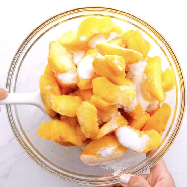 Peaches being tossed with sugar in a large glass bowl.