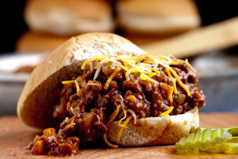 A close-up of a sloppy joe sandwich filled with saucy, seasoned beef, topped with shredded cheese on a bun. In the foreground, thinly sliced pickles are visible, and in the background, blurred buns are stacked.